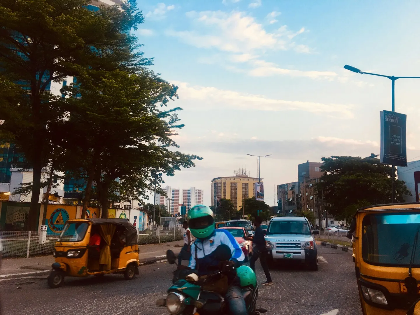 A rider in a green helmet on a motorbike in Lagos traffic, between yellow tricycles, with office towers behind.